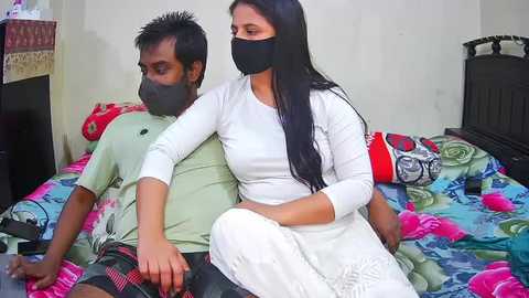 Media: Video of a South Asian couple on a bed, the man in a green shirt and the woman in white clothes, both masked, in a modest bedroom with colorful floral bedding.