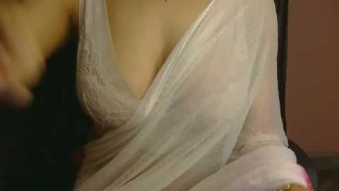 A close-up video of a woman wearing a sheer, white, lace-trimmed gown, revealing her cleavage and partially see-through fabric, with blurred background suggesting an intimate indoor setting.