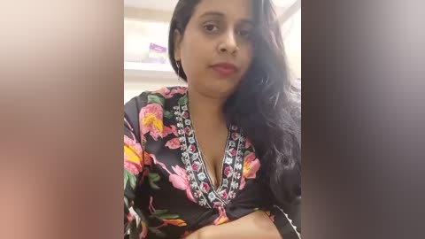 Media: Video of a South Asian woman with medium skin tone and long black hair, wearing a black floral-patterned blouse with a deep V-neck. She has a neutral expression and is indoors, likely in a bathroom with a white shelf visible in the blurred background.