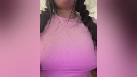 A video of a woman with light skin, wearing a tight, light pink ribbed top that accentuates her ample breasts. She has long, black hair in a single braid. The background is blurred, featuring a pink gradient effect.