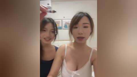 Video of two smiling Asian women with fair skin, one in a blue dress, the other in a white camisole, standing in a well-lit room with framed art on the wall.