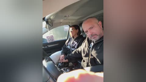 Media: Video of two men, one bald, wearing black jackets, driving a car with a green van visible through the window. The lighting is bright, suggesting daytime.