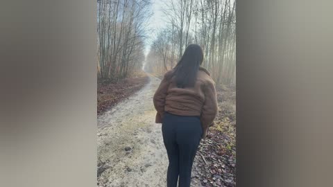 A video captures a lone woman walking away from the camera, dressed in a brown coat and blue jeans, on a foggy, leaf-strewn forest path.