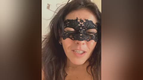 Video of a woman with long dark hair, wearing a black lace masquerade mask. She has a fair complexion and is smiling slightly. The background is blurred.