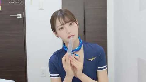 Video of an Asian woman in a blue flight attendant uniform, holding a clear tube to her mouth, indoors with white walls and dark wood doors.