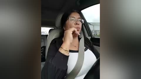 Video of a woman with long black hair, wearing glasses and a black shirt, seated in a car with seatbelt on.