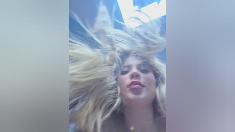 Video of a blonde woman with long, tousled hair, wearing a white top, captured from a low angle, with a blurred, blue background.