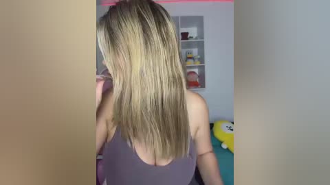 Video of a blonde woman with straight hair, wearing a purple tank top, standing in a colorful room with shelves, plush toys, and a yellow character.