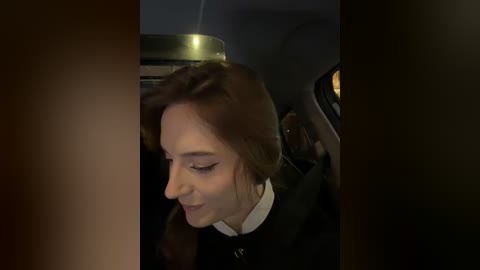 Video of a young woman with long brown hair, wearing a black jacket, leaning forward in a car with dim interior lighting.