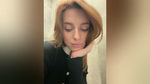 Video of a fair-skinned woman with long, straight red hair, wearing a black coat, looking down thoughtfully. The background is blurred, creating a soft focus effect.