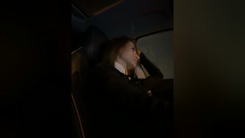 Video of a young woman with long brown hair, dressed in a black suit, seated in a dimly lit car, looking out the window, deep in thought.