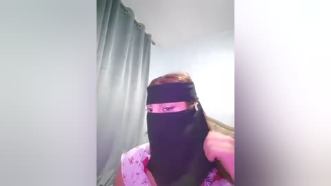 A video of a person wearing a black face mask and a light pink shirt with white polka dots, standing in a room with gray curtains. The image is slightly out of focus.