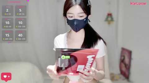 Video of an Asian woman with long black hair, wearing a face mask and headphones, reading a book in a white room.