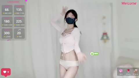 Video of an East Asian woman with pale skin, wearing a pink crop top, white panties, and a black face mask, posing in a white room.
