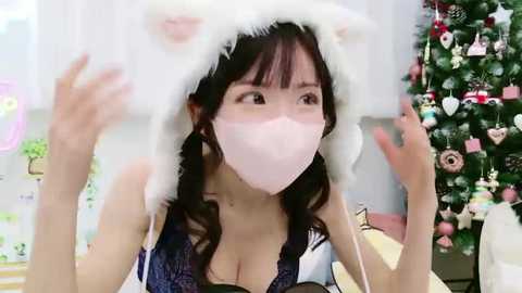 Video of an East Asian woman with long dark hair in a cozy room. She wears a white cat hat, pink mask, and a low-cut blue dress. Background includes a decorated Christmas tree and soft lighting.