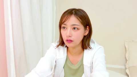 A video of an East Asian woman with shoulder-length brown hair, wearing a white lab coat over a green top, sitting in a clinical setting with beige walls and a white curtain.
