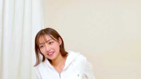 Video of a young Asian woman with shoulder-length brown hair, wearing a white hoodie, smiling in a minimalistic, indoor setting with beige walls and white curtains.