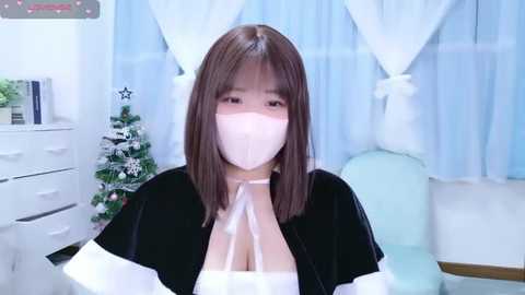 Video of a young Asian woman with straight, shoulder-length brown hair, wearing a black velvet cape over a white dress and a pink face mask, standing in a white room with a Christmas tree and blue curtains.
