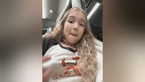 A video of a young woman with curly blonde hair and fair skin wearing a white T-shirt featuring a cartoon, sitting in a car with a blurred background.
