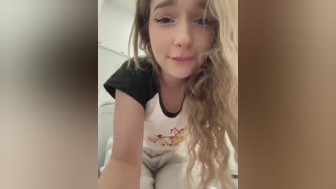 Video of a young woman with long, wavy blonde hair, wearing a black and white graphic T-shirt, sitting on a bed with a beige blanket. She has a neutral expression and is indoors with a blurred background.