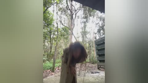 A video of a blurred, faceless person in a brown coat standing in a forest, surrounded by tall trees and a green utility trailer. The background is overexposed, creating a hazy effect.