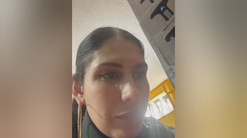 A close-up video of a woman with dark hair, wearing a black top, standing in front of a yellow wall with a white ceiling fan, and a window with greenery visible in the background.