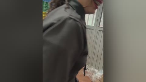Video of a woman with medium brown skin and curly hair, wearing a black leather jacket, standing in a hallway with a gray door and colorful shelves in the background.
