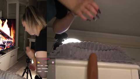 Video of a blonde woman in a black top and shorts, recording herself on a tripod-mounted camera, in a dimly lit room with a TV and bed visible.