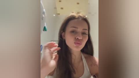 Video of a young woman with long brown hair, wearing a white tank top, blowing a kiss into a mirror. Background shows a tiled wall and a decorative light fixture.