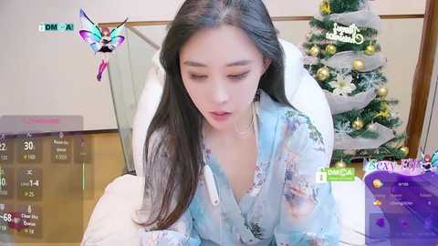 Video of an East Asian woman with long black hair, wearing a light blue floral robe, leaning forward on a bed. Christmas tree with ornaments in background.