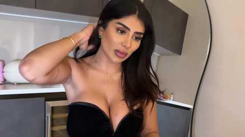 Video of a curvy Latina woman with long dark hair, large breasts, and full lips, wearing a black strapless velvet dress, standing in a modern kitchen.