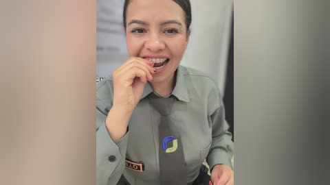Video of a smiling Latina woman with dark hair, braces, and a gray uniform with a logo, standing in a hallway.