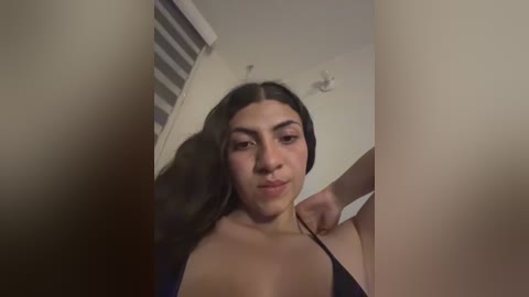 Video of a Latina woman with long, dark hair, medium skin tone, and medium-sized breasts, wearing a black bra. She stands in a dimly lit room with a white wall and blinds partially covering a window.