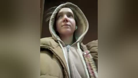 A video of a young person with pale skin and light brown hair, wearing a beige puffer jacket and a light-colored hoodie, looking up thoughtfully. The background is blurred and dark.