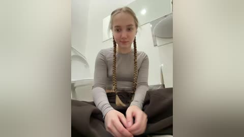 Video of a young woman with light skin and long braided hair, wearing a grey long-sleeve top, sitting on a dark carpeted floor, looking slightly downward, in a modern, clean bathroom with white walls and a mirror.