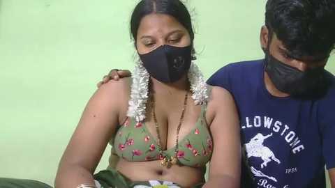 Video of a South Asian woman with medium-dark skin, wearing a floral green bra, white dhoti, and face mask, sitting beside a man with black beard and dark skin in a blue shirt.
