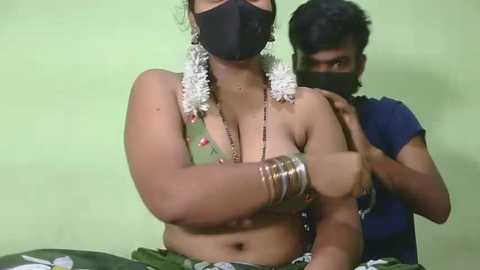 Video of an Indian woman with medium brown skin, wearing a black face mask, traditional green blouse, white flowers, and multiple bangles, standing in front of a man in a blue t-shirt, both indoors against a light green wall.