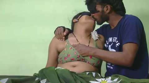 Video of an intimate moment between a dark-skinned man and a fair-skinned woman, both wearing casual clothing, kissing passionately. The woman is lying on a green bedspread, her hair tied back.