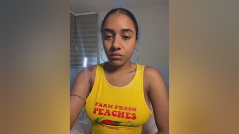 Video of a young woman with medium brown skin, wearing a yellow \"Farm Fresh Peaches\" tank top and hoop earrings, standing indoors.