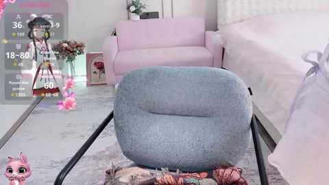 A video of a chic, minimalist bedroom with light pink furniture, featuring a cozy, textured blue armchair, a white bed, and a soft grey carpet. A virtual doll stands in the corner.