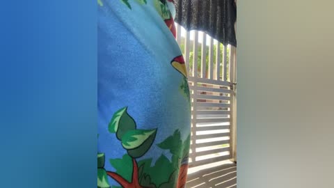 A video of a plush toy parrot perched on a window ledge, partially obscured by green leafy plants, with sunlight streaming through vertical blinds. The toy's vibrant colors contrast against the muted background.
