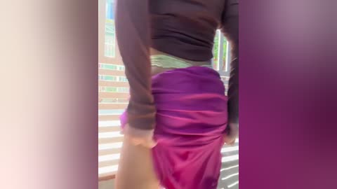A video captures a person in a dark shirt and a bright purple skirt, standing in front of a window with vertical blinds. The image is slightly blurry, creating a soft, dreamy effect.