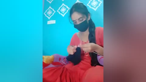 Video of a woman in a red dress and black mask sewing a black garment on a couch, teal wall with geometric patterns, blurred background.