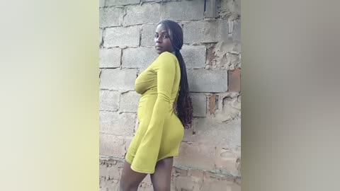 Media: Video of a curvaceous Black woman with dark skin, wearing a bright yellow, long-sleeved, high-waisted mini dress, posing against a textured, beige brick wall.