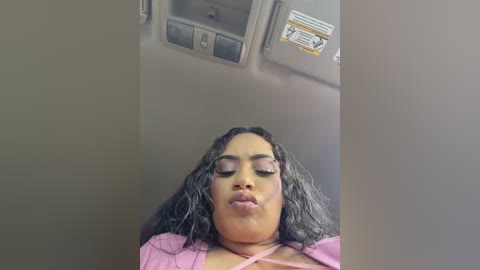 Video of a young Black woman with long, curly black hair and fair skin, wearing a pink top, puckering her lips in a suggestive pose inside an airplane's overhead compartment.