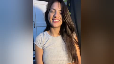 Video of a smiling woman with long dark hair, wearing a light grey ribbed crop top, standing indoors with white door and grey wall in the background.