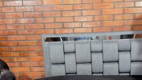 Video of a modern interior featuring a grey, tufted leather headrest with a grid pattern against a brick wall. The brick wall has a rustic, earthy tone with a slight reddish hue. The foreground shows a dark wooden table.