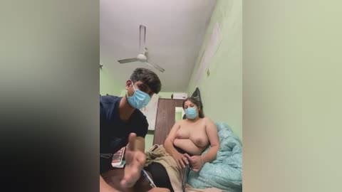 Video of a man wearing a blue surgical mask and a woman with a blue mask and bare breasts, sitting in a green-painted room with a ceiling fan.