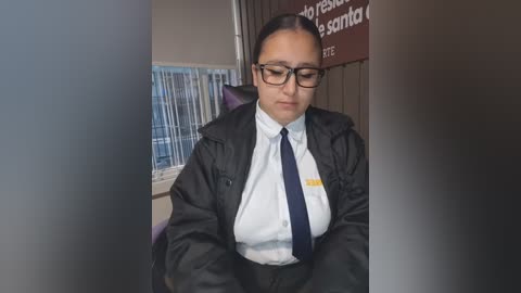 Media: Video of a Latina woman with dark hair, wearing glasses, a black jacket, white shirt, and dark tie, seated at a desk with a window and blinds in the background.