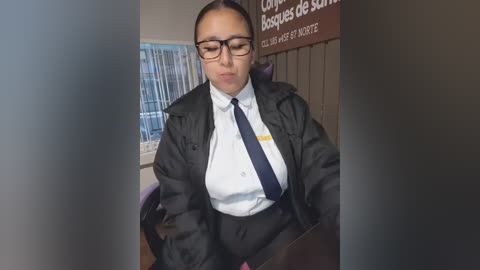Video of a woman in a dark jacket over a white shirt and tie, sitting at a desk in an office with a French sign and window blinds.